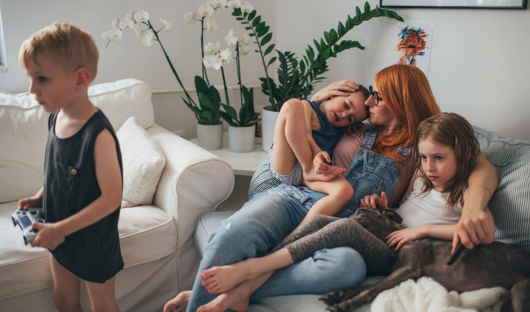 mother sits on a couch with her daughters while her son plays a video game